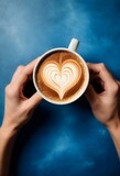 A pair of hands holding a cup of coffee with heart-shaped latte art on a blue textured background with steam rising from the cup.