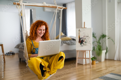 Fotografie Beautiful artsy girl, sitting in modern, bright, nicely decorated living room, working on her laptop and smiling while reading positive customer feedback