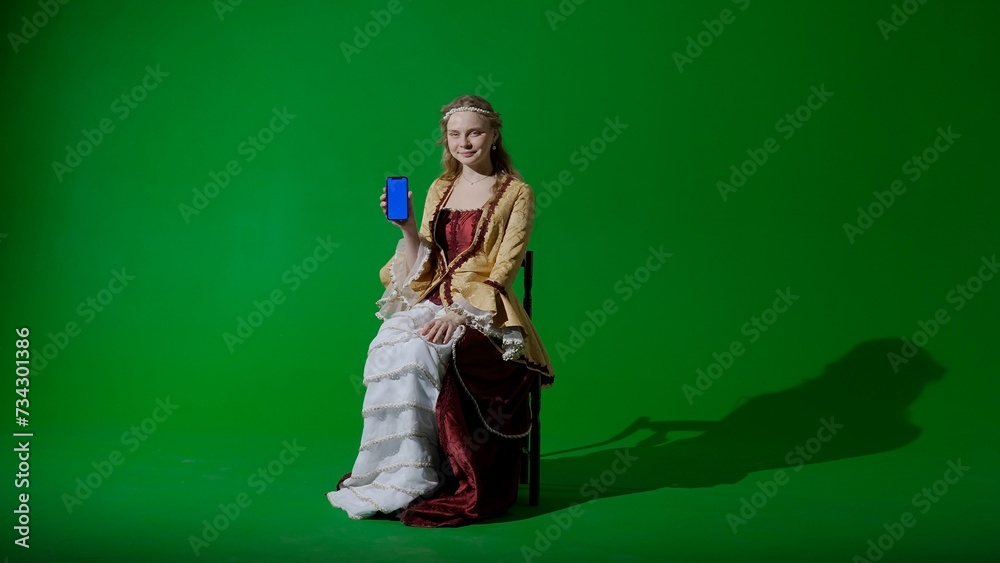 Woman in ancient outfit on chroma key green screen background. Female in renaissance dress holding smartphone with workspace mockup, shows thumb up.