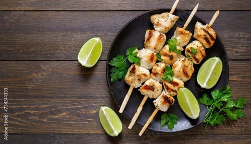 Grilled chicken souvlaki with parsley and lime wedges on black plate on dark wooden table, horizontal view from above, flat lay, free space photo