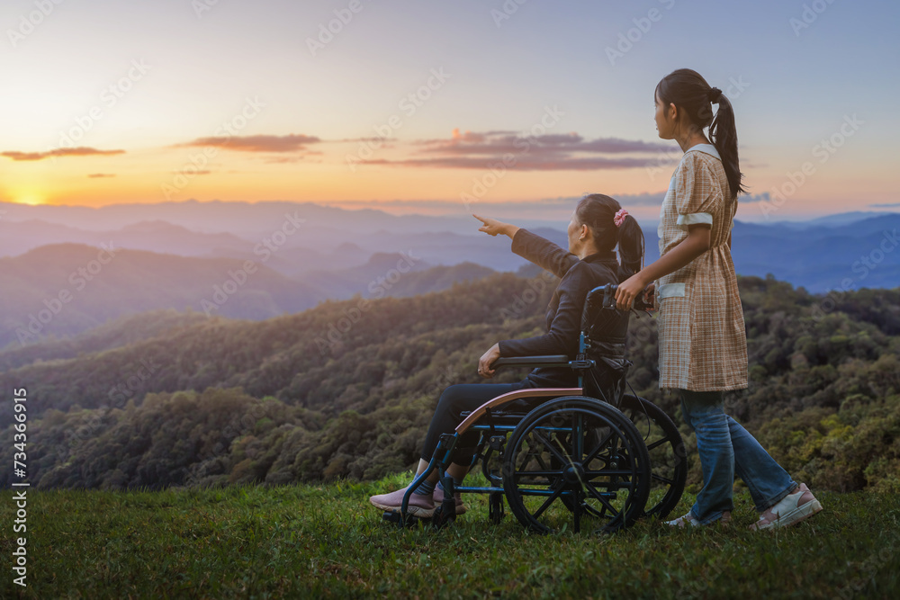 Woman sitting in wheelchair walking with his care helper on mountain sunset .Silhouette