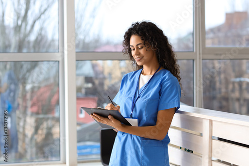 Smiling female nurse in blue scrubs writing on clipboard at hospital receptio...
