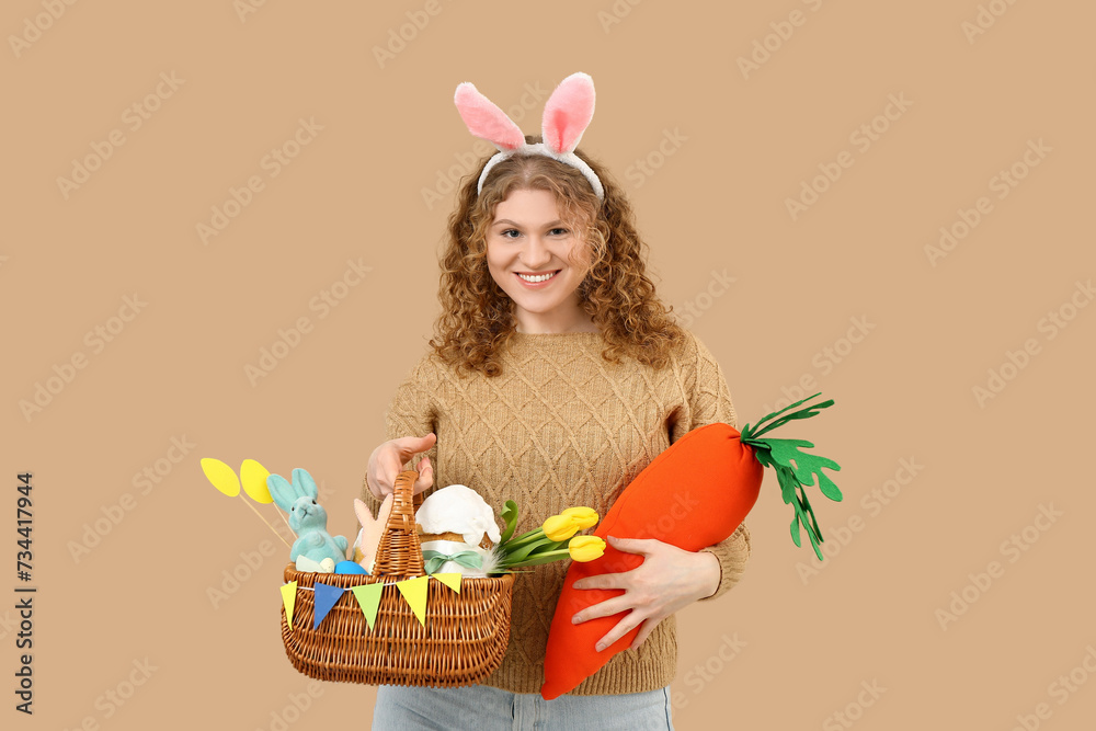 Beautiful young happy woman in bunny ears with carrot-shaped toy and Easter basket on beige background