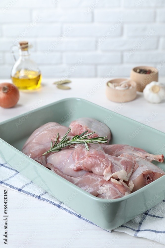 Raw rabbit meat and rosemary in baking dish on white wooden table, closeup