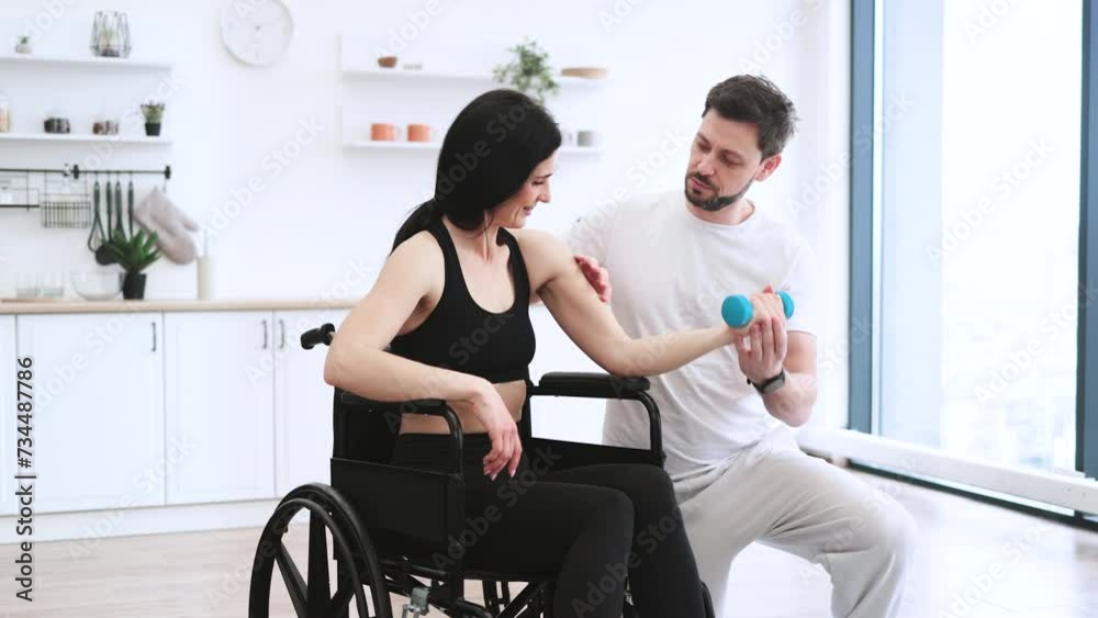 Handicapped mature woman training with dumbbells. Physiotherapist or husband in sportswear helping Caucasian female patient in wheelchair exercise at home. Rehabilitation of disabled people concept. Handicapped mature woman training with dumbbells. Physiotherapist or husband in sportswear helping Caucasian female patient in wheelchair exercise at home. Rehabilitation of disabled people concept.