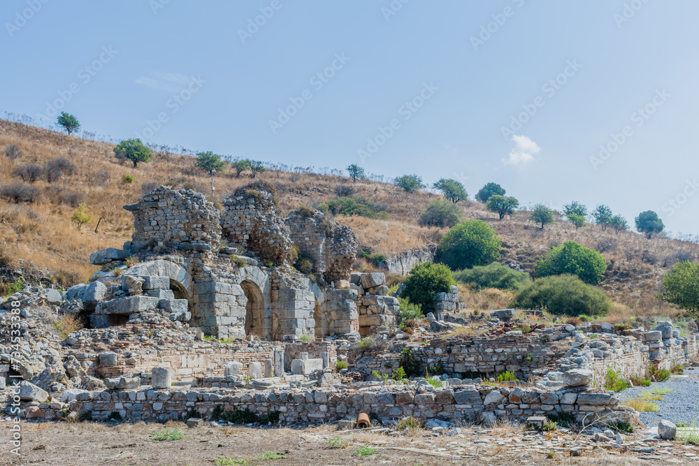 Ruins of stone arches against a clear sky with sparse greenery in Ephesus, Turkiye.