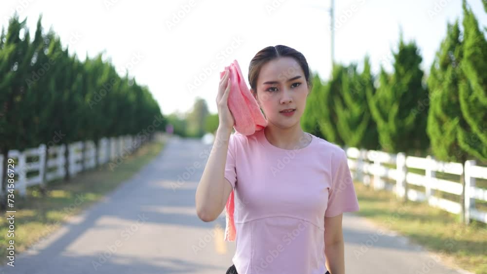 A beautiful healthy Asian woman uses a towel to wipe away her sweat while taking a walk to rest from jogging. A beautiful healthy Asian woman uses a towel to wipe away her sweat while taking a walk to rest from jogging.