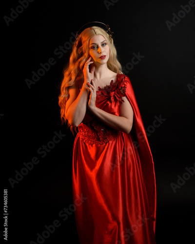 Close up portrait of beautiful blonde model wearing flowing red silk toga gown and crown, dressed as ancient mythological fantasy goddess. Graceful elegant pose isolated on dark studio background.