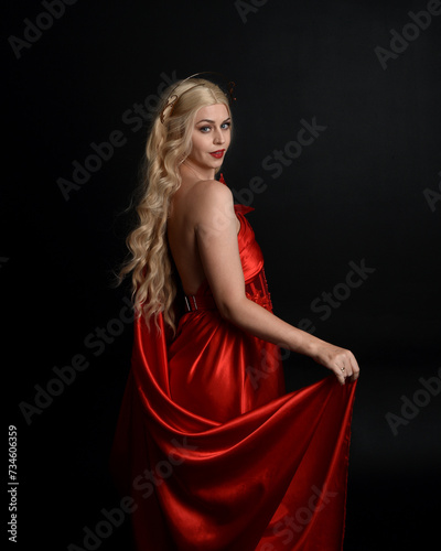 Close up portrait of beautiful blonde model wearing flowing red silk toga gown and crown, dressed as ancient mythological fantasy goddess. Graceful elegant pose isolated on dark studio background.