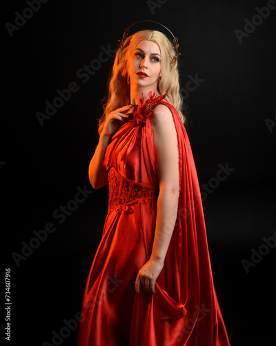 Close up portrait of beautiful blonde model wearing flowing red silk toga gown and crown, dressed as ancient mythological fantasy goddess. Graceful elegant pose isolated on dark studio background.