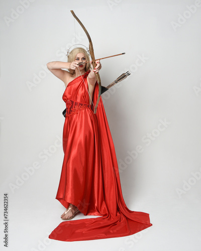 Full length portrait of blonde model dressed as mythological fantasy goddess in flowing red silk toga gown, crown. Graceful elegant pose holding archery weapons, isolated on studio background