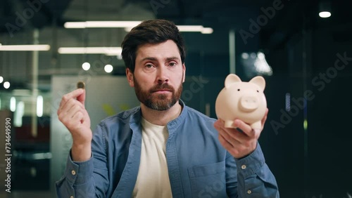 Unassured caucasian man looking at coin and classic piggy bank while raising hands in uncertainty. Adult guy in casual outfit thinking about investing for getting passive income from banking deposit. photo