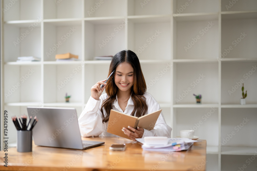 A cheerful young businesswoman writing notes with a pen in a notebook at her organized workspace with a laptop A cheerful young businesswoman writing notes with a pen in a notebook at her organized workspace with a laptop