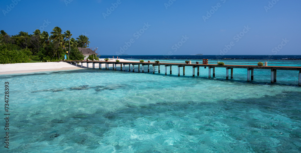 Wooden bridge over the sea leading to an island with sandy beaches and a green forest. Palm trees.