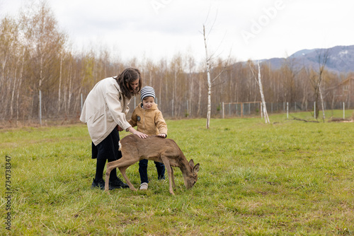 Mother and Child Petting a Deer On The Grass. Outdoor Family Activities © Doralin