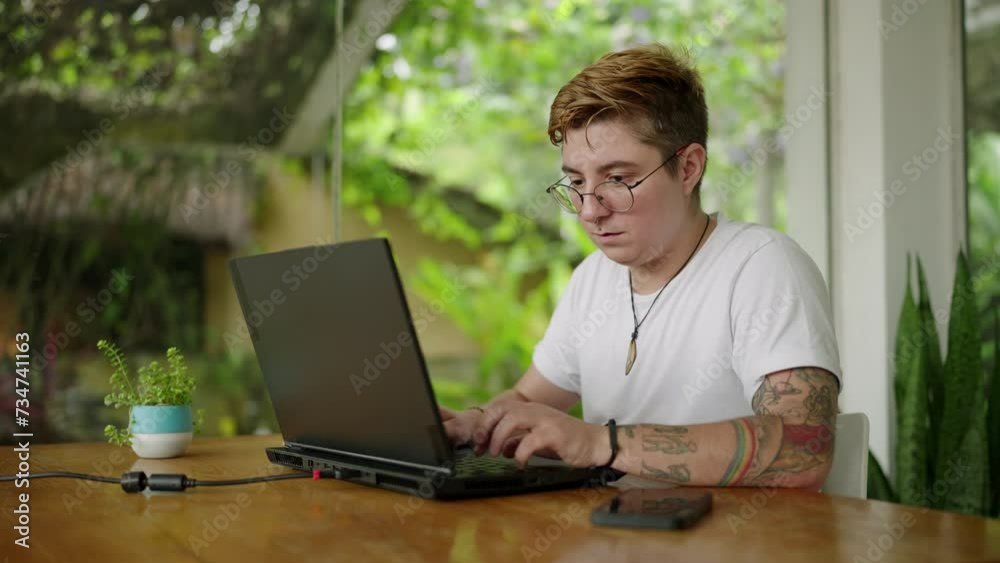 Tattooed individual in glasses progresses work on laptop, serene open-plan office setting. Inclusive, modern workspace vibe. Pro engrossed in task, amidst green plants, daylight floods area. Slowmo Tattooed individual in glasses progresses work on laptop, serene open-plan office setting. Inclusive, modern workspace vibe. Pro engrossed in task, amidst green plants, daylight floods area. Slowmo