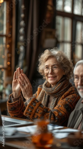 An elderly woman wearing glasses and a scarf smiles and raises her hand in a room
