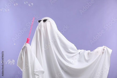 Funny ghost. Person in white sheet and sunglasses blowing soap bubbles on violet background