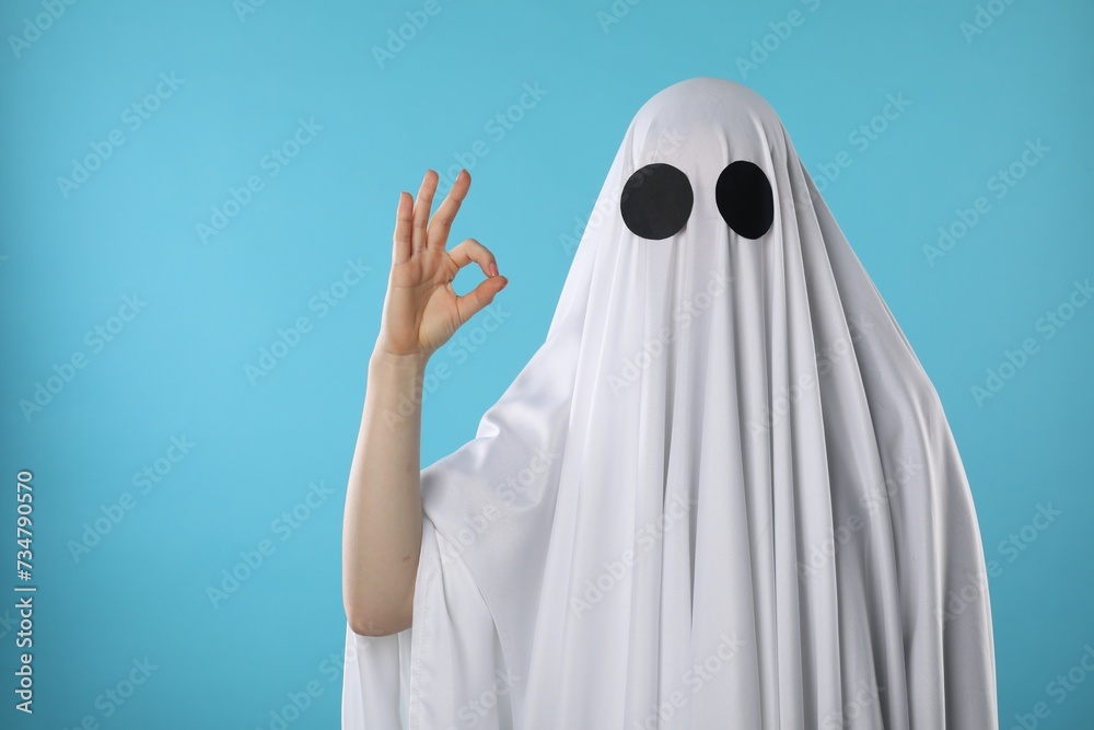 Funny ghost. Woman covered with white sheet showing OK gesture on light blue background