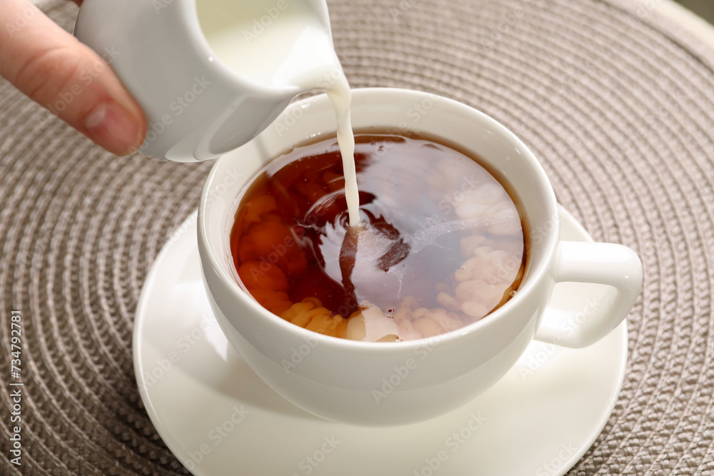 Woman pouring milk into cup with aromatic tea at table, closeup Woman pouring milk into cup with aromatic tea at table, closeup