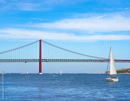 Motorboats, container ship, and sailboats sailing on the Tagus River with the red steel 25 de Abril suspension bridge in the background with road traffic accessing Lisbon, under a clear blue sky.
