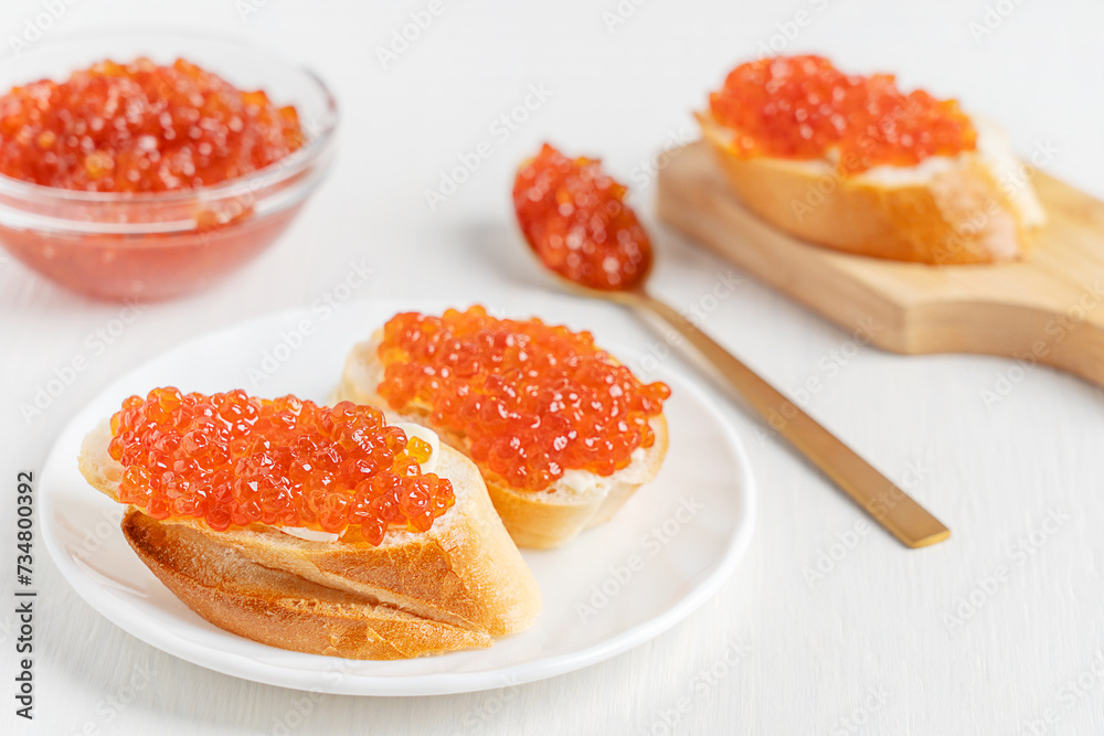 Sliced loaf of bread or canape with butter spread and salted organic salmon fish roe or red caviar served on plate on white wooden table with spoon, chopping board and glass bowl as healthy snack Sliced loaf of bread or canape with butter spread and salted organic salmon fish roe or red caviar served on plate on white wooden table with spoon, chopping board and glass bowl as healthy snack