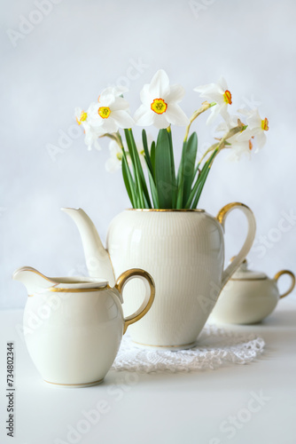 Still life with a blooming bouquet of white daffodils in an elegant porcelain teapot on a textured white background