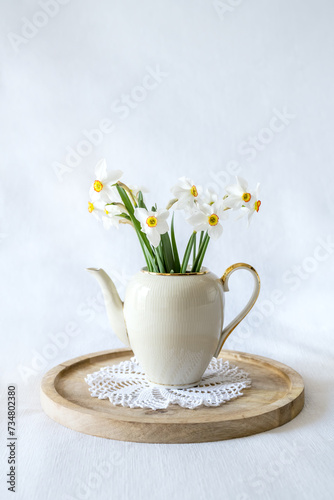 Still life with a blooming bouquet of white daffodils in an elegant porcelain teapot on a textured white background
