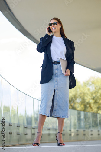 Confident fashionable young woman dressed in an elegant look of a skirt and jacket speaks on the phone while walking down the street © makedonski2015