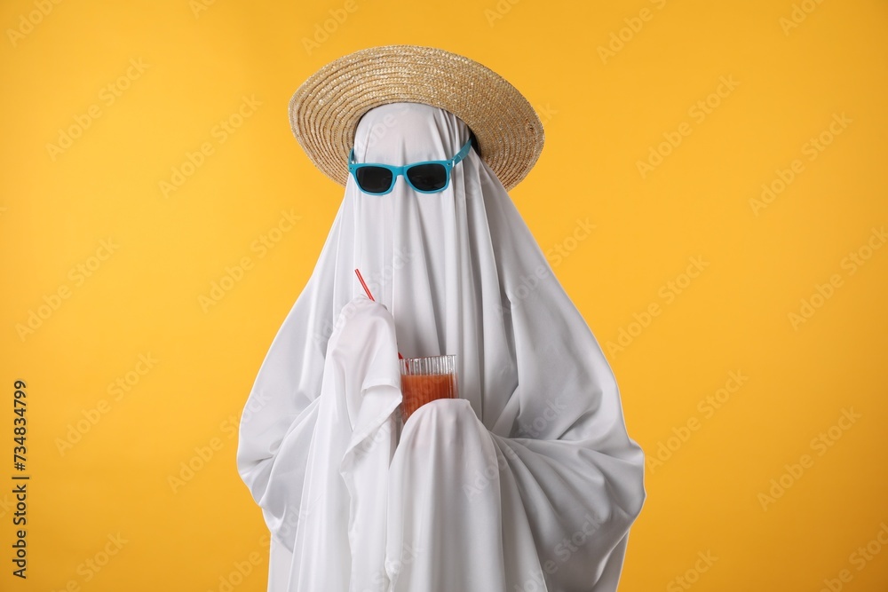 Person in ghost costume, sunglasses and straw hat holding glass of drink on yellow background