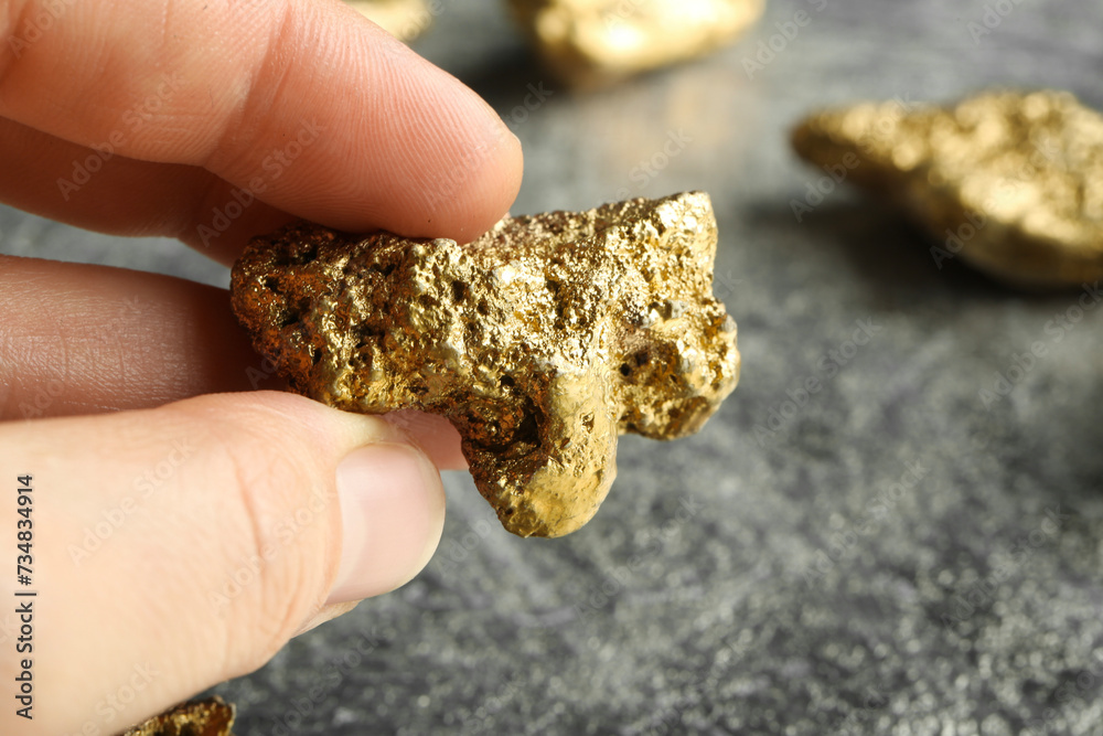 Woman holding gold nugget at grey textured table, closeup. Space for text Woman holding gold nugget at grey textured table, closeup. Space for text