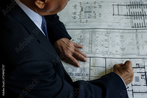 The picture shows a male engineer in a suit working with electrical network drawings, at an engineer's workplace.