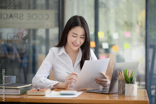 Happy asian young businesswoman holding documents folders in office working space, Asian female employee using laptop talking on the phone at workplace.