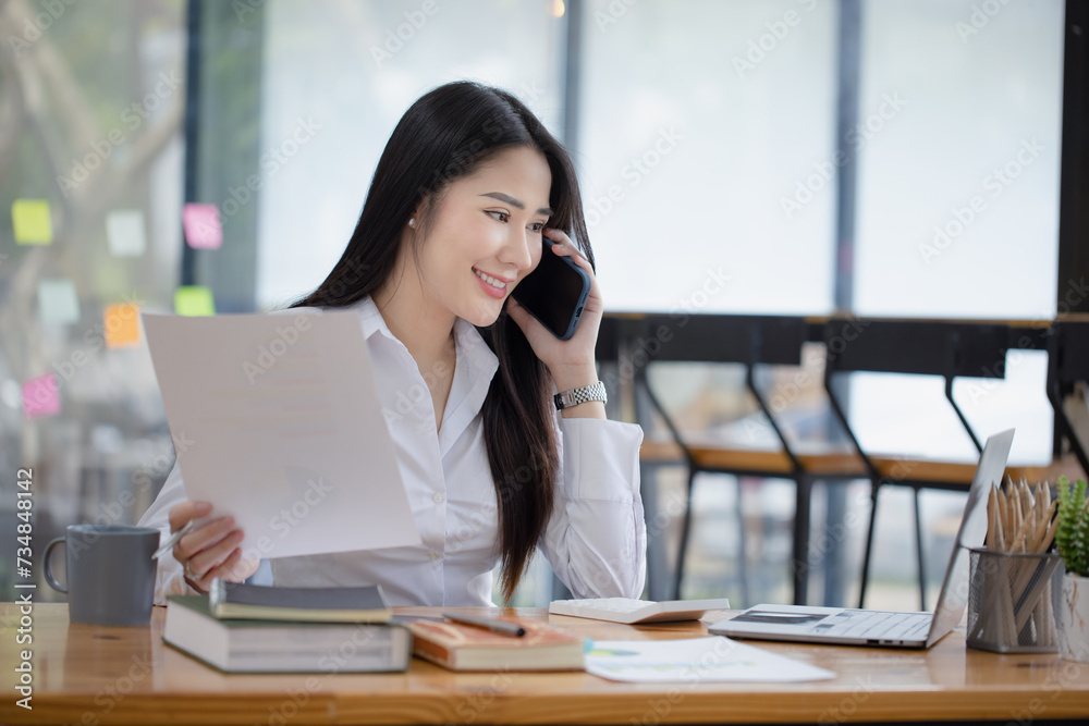 Happy asian young businesswoman using digital smartphone sitting in office working space, Asian female employee using laptop talking on the phone at workplace. Happy asian young businesswoman using digital smartphone sitting in office working space, Asian female employee using laptop talking on the phone at workplace.