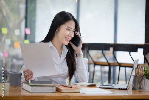 Happy asian young businesswoman using digital smartphone sitting in office working space, Asian female employee using laptop talking on the phone at workplace. © SOMKID