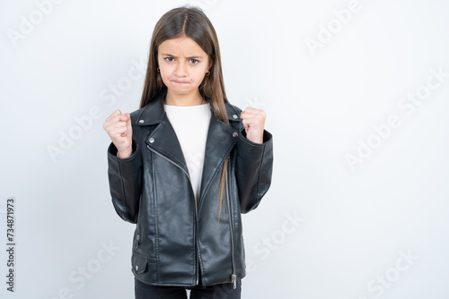 Foto Irritated Young beautiful teen girl wearing biker jacket blows cheeks with anger and raises clenched fists expresses rage and aggressive emotions