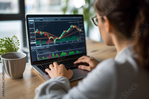 Caucasian woman in glasses sitting at the table in front of laptop with stock market chart - rear over shoulder view. © lucky pics