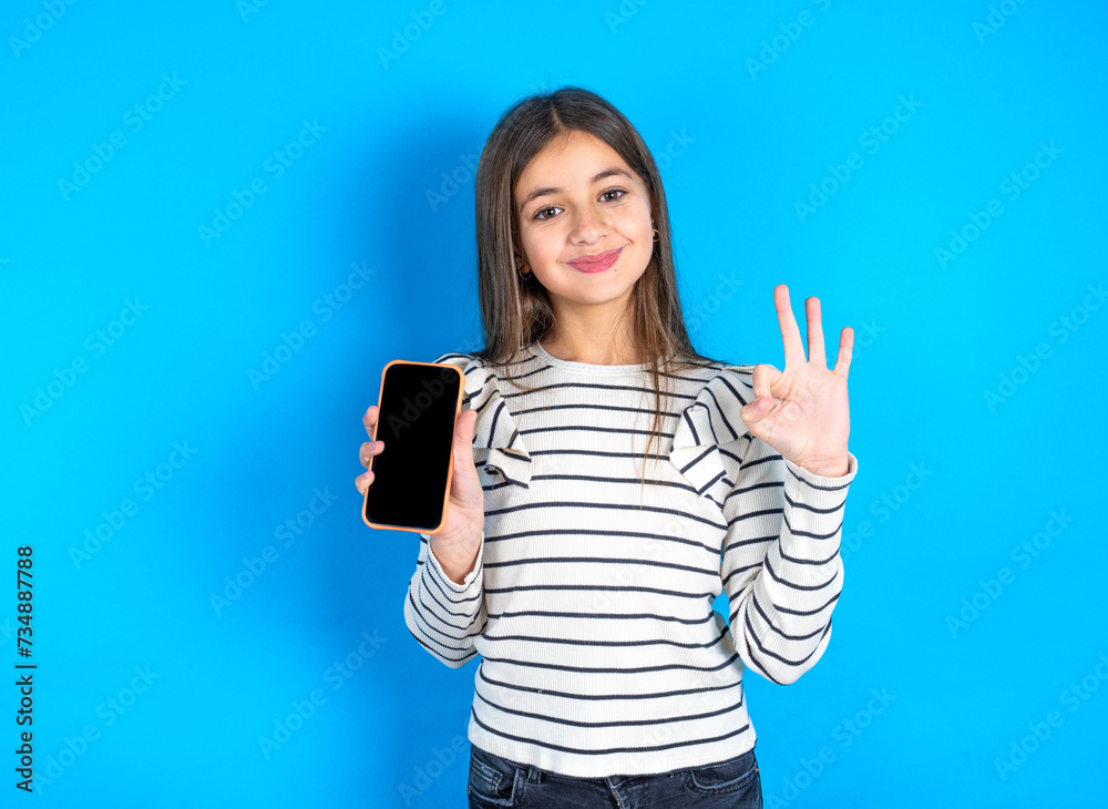 Beautiful kid girl wearing striped T-shirt holding in hands cell showing ok-sign Beautiful kid girl wearing striped T-shirt holding in hands cell showing ok-sign