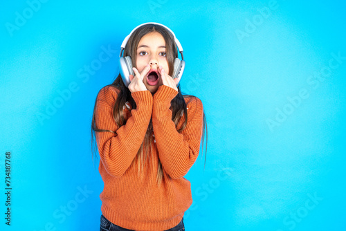 Shocked beautiful kid girl wearing orange sweater stares fearful at camera keeps mouth widely opened wears wireless stereo headphones on ears photo