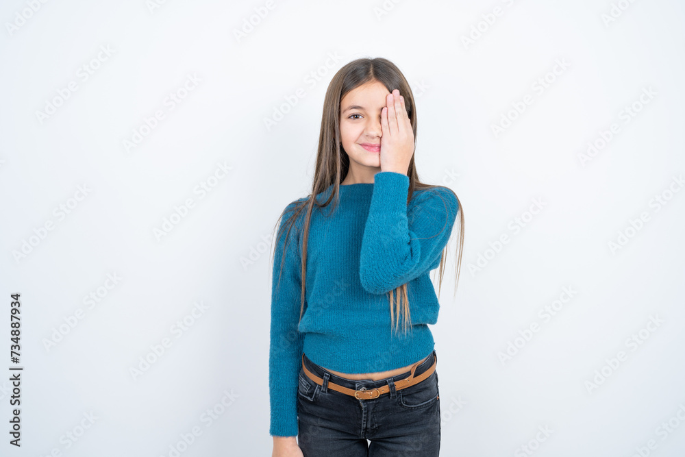 beautiful kid girl wearing blue sweater covering one eye with her hand, confident smile on face and surprise emotion. beautiful kid girl wearing blue sweater covering one eye with her hand, confident smile on face and surprise emotion.