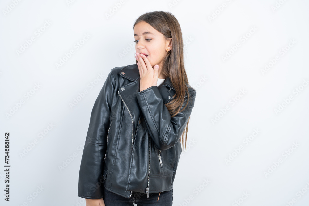 beautiful kid girl wearing biker jacket covers mouth and looks with wonder at camera, cannot believe unexpected rumors. beautiful kid girl wearing biker jacket covers mouth and looks with wonder at camera, cannot believe unexpected rumors.