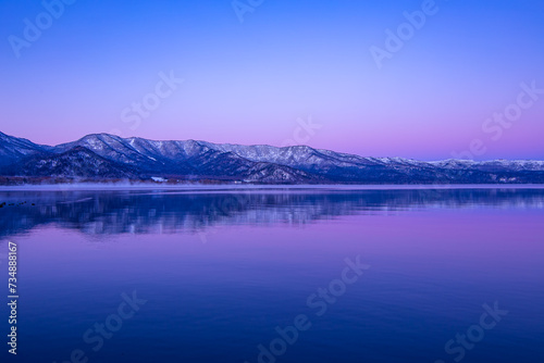 淡い紫色のグラデーションに染まる夜明けの空と雪山を水面に反射する湖。日本の北海道の旅行地である屈斜路湖。雄大で美しい自然。 © Masa Tsuchiya