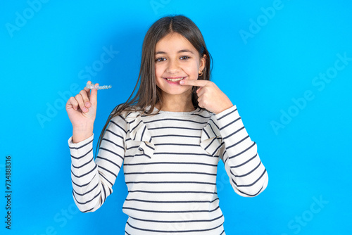 beautiful kid girl wearing  striped T-shirt holding an invisible aligner and pointing to her perfect straight teeth. Dental healthcare and confidence concept.