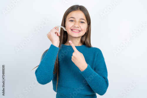 beautiful kid girl wearing blue sweater holding an invisible aligner and pointing at it. Dental healthcare and confidence concept.