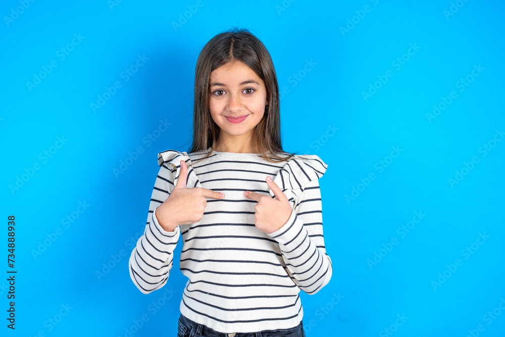 Fototapeta premium Embarrassed Young kid girl wearing striped t-shirt indicates at herself with puzzled expression, being shocked to be chosen to participate in competition, hesitates about something
