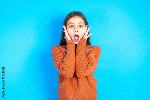 Happy Young kid girl wearing orange knitted sweater touches both cheeks gently, has tender smile, shows white teeth, gazes positively straightly at camera, photo