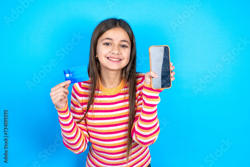 Young kid girl wearing striped t-shirt opened bank account, holding smartphone and credit card, smiling, recommend use online shopping application