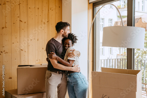 Happy couple embracing each other while standing near window at new home photo