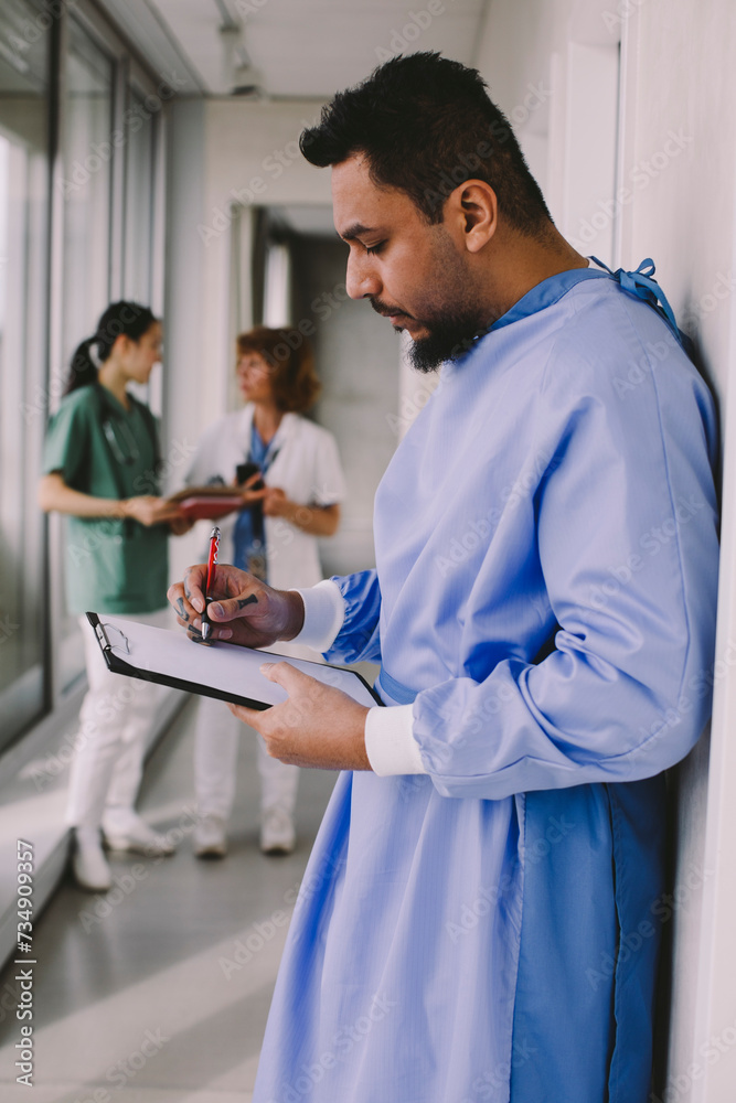 Side view of male surgeon writing on clipboard in hospital corridor Side view of male surgeon writing on clipboard in hospital corridor