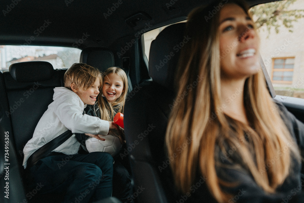 Happy brother and sisters sitting behind while mother driving car on weekend Happy brother and sisters sitting behind while mother driving car on weekend